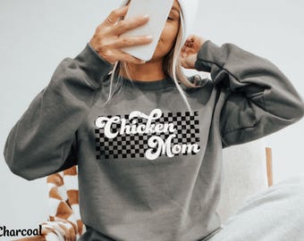 May include: Charcoal gray sweatshirt with the text "Chicken Mom" in white script over a black and white checkered design. The sweatshirt has long sleeves and a crew neck. The word "Charcoal" is printed at the bottom left.