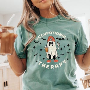 May include: Teal t-shirt with the words "OCCUPATIONAL THERAPY" surrounding a cartoon ghost wearing a red hat and holding a drink. The ghost is giving a peace sign. Bats and diamonds are also in the design.