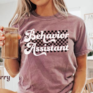 May include: Berry-colored t-shirt with the words "Behavior Assistant" in a white, retro font over a black and white checkered background. The person is holding a glass jar with a straw and a brown liquid.