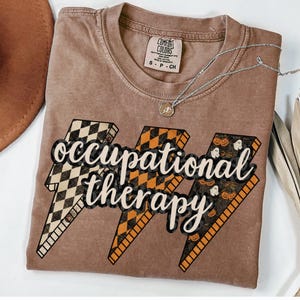 May include: Brown t-shirt with the words "occupational therapy" in a stylized font, set against three lightning bolt graphics. The lightning bolts feature checkered, orange, and Halloween-themed patterns. A silver necklace with a pendant is draped across the shirt.