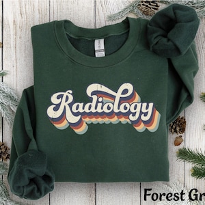 May include: Forest green sweatshirt with the word "Radiology" in a retro font. The word is in white with a rainbow design. The sweatshirt is folded on a wooden surface with pine needles and cones.