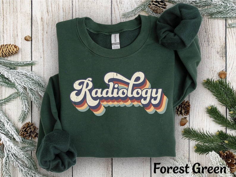 May include: Forest green sweatshirt with the word "Radiology" in a retro font. The word is in white with a rainbow design. The sweatshirt is folded on a wooden surface with pine needles and cones.