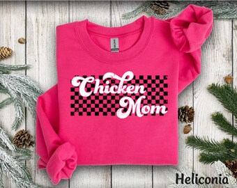May include: A bright pink sweatshirt with the words "Chicken Mom" in white, stylized script over a black and white checkered pattern. The sweatshirt is a crew neck style, with the sleeves folded. The background includes pinecones and greenery.