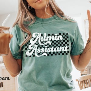 May include: Seafoam green t-shirt with the words "Admin Assistant" in white script over a black and white checkered pattern. The shirt is a casual style, suitable for everyday wear.