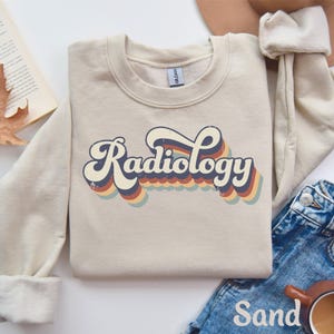 May include: A sand-colored sweatshirt with the word "Radiology" in a retro font. The text has a layered design with blue, red, and yellow accents. The sweatshirt is paired with blue jeans and a brown hat.