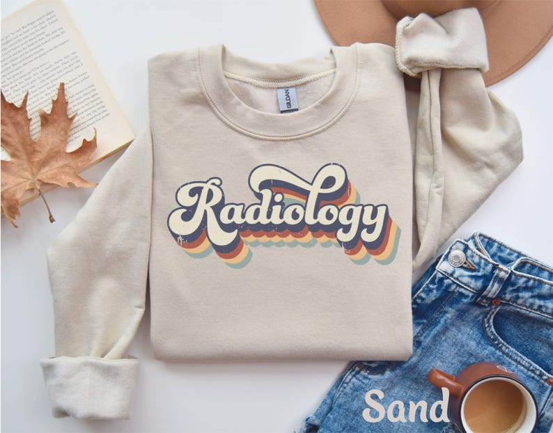 May include: A sand-colored sweatshirt with the word "Radiology" in a retro font. The text has a layered design with blue, red, and yellow accents. The sweatshirt is paired with blue jeans and a brown hat.