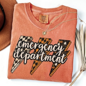 May include: Coral-colored t-shirt with the words "emergency department" in a stylized font, set against three lightning bolt graphics. The lightning bolts feature checkered, orange, and Halloween-themed patterns. A gold necklace and a brown hat are also visible.