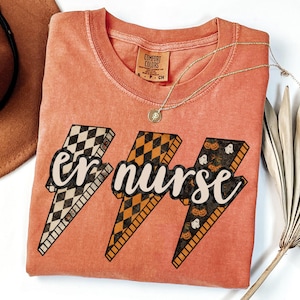 May include: Coral-colored t-shirt with the text "er nurse" in a lightning bolt design. The lightning bolts feature checkered, orange diamond, and Halloween-themed patterns. A gold necklace and a brown hat are also visible.