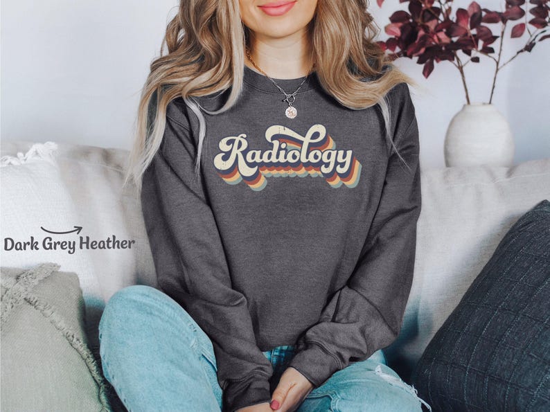 May include: Dark grey heather sweatshirt with the word "Radiology" in a retro font. The word is in a cream color with a rainbow-colored wavy design underneath. The sweatshirt is a crew neck style.