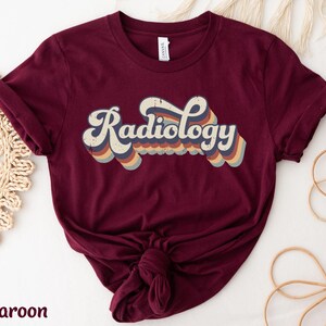 May include: Maroon t-shirt with the word "Radiology" in a retro font. The text is in white with a layered design of blue, orange, and yellow stripes. The shirt is knotted at the bottom.
