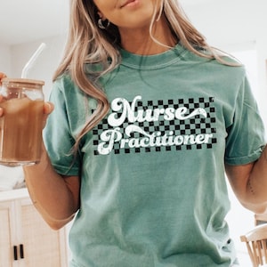 May include: A teal-colored t-shirt with the words "Nurse Practitioner" in a white, retro font over a black and white checkered design. The shirt is short-sleeved and appears to be made of a soft material.