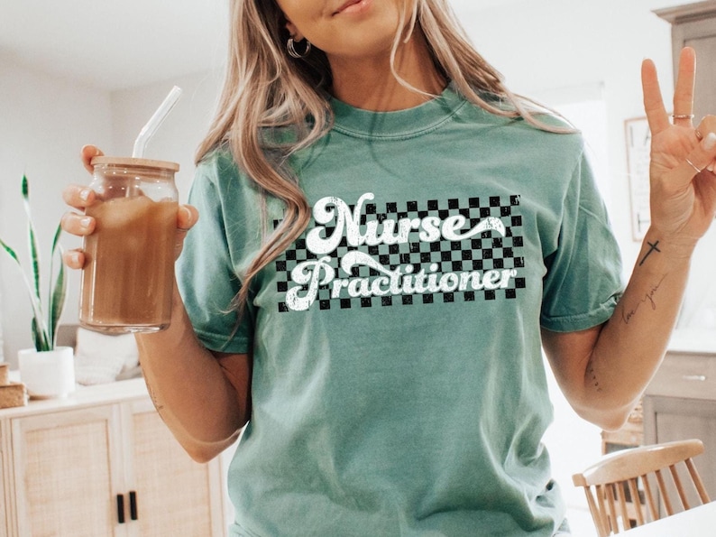 May include: A teal-colored t-shirt with the words "Nurse Practitioner" in a white, retro font over a black and white checkered design. The shirt is short-sleeved and appears to be made of a soft material.