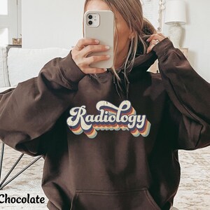 May include: Dark chocolate-colored hoodie with the word "Radiology" in a retro font across the chest. The text has a layered design with red, orange, yellow, and blue accents. The hoodie has a front pocket and a hood.