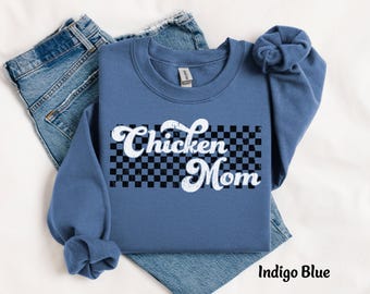 May include: Indigo blue sweatshirt with the text "Chicken Mom" in a white, retro font over a black and white checkered pattern. The sweatshirt is folded over a pair of blue jeans.