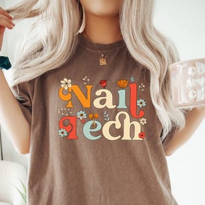 K&ouml;nnte beinhalten: Braunes T-Shirt mit dem Schriftzug "Nail Tech" in einer Retro-Schriftart, verziert mit Blumen. Das Shirt hat eine neutrale Farbe und eine Vintage-&Auml;sthetik. Eine rosa Tasse mit den Worten "Cool Mom Club" wird in der rechten Hand gehalten.