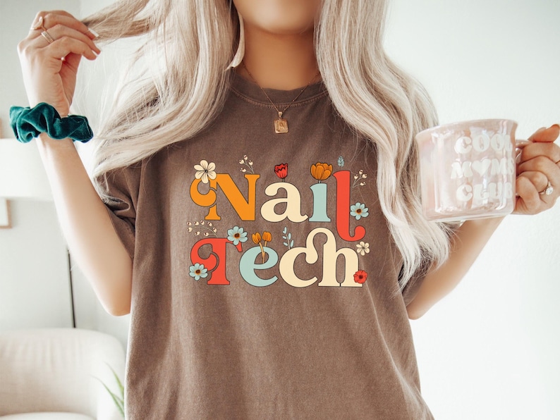 K&ouml;nnte beinhalten: Braunes T-Shirt mit dem Schriftzug "Nail Tech" in einer Retro-Schriftart, verziert mit Blumen. Das Shirt hat eine neutrale Farbe und eine Vintage-&Auml;sthetik. Eine rosa Tasse mit den Worten "Cool Mom Club" wird in der rechten Hand gehalten.