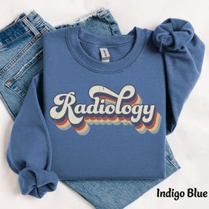 May include: Indigo blue sweatshirt with the word "Radiology" in a retro font. The word is in white with a multi-colored wave design underneath. The sweatshirt is folded on top of a pair of blue jeans.