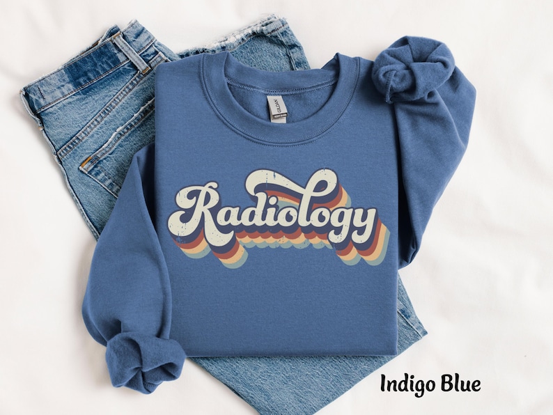 May include: Indigo blue sweatshirt with the word "Radiology" in a retro font. The word is in white with a multi-colored wave design underneath. The sweatshirt is folded on top of a pair of blue jeans.