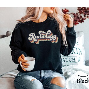 May include: Black sweatshirt with the word "Radiology" in a retro font. The text is in white with a rainbow-colored outline. The sweatshirt is a crew neck style. The image also shows a person holding a white mug.