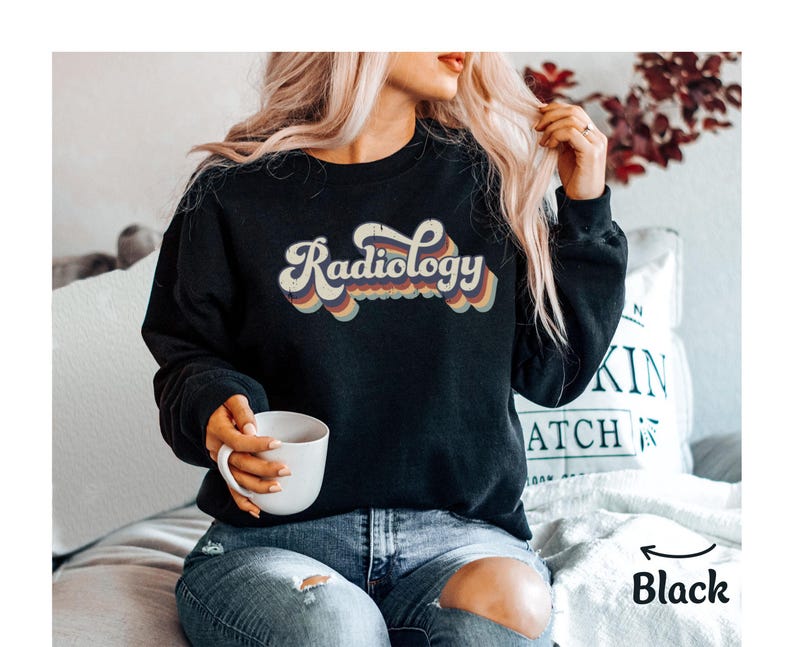 May include: Black sweatshirt with the word "Radiology" in a retro font. The text is in white with a rainbow-colored outline. The sweatshirt is a crew neck style. The image also shows a person holding a white mug.