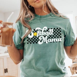 May include: A teal green t-shirt with a vintage-style graphic that says "Softball Mama" with a checkered background and a yellow softball.