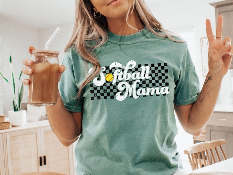 May include: A teal green t-shirt with a vintage-style graphic that says "Softball Mama" with a checkered background and a yellow softball.