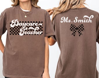 May include: Two brown t-shirts with a checkered pattern and text. The front of one shirt says "Daycare Teacher" and the back of the other shirt says "Ms. Smith" with a checkered bow.