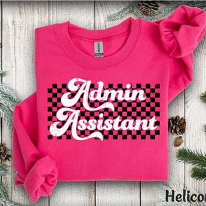 May include: A bright pink sweatshirt with the words "Admin Assistant" in white script over a black and pink checkered pattern. The sweatshirt is displayed on a wooden surface with decorative elements.