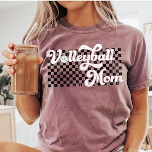 Puede incluir: Una camiseta malva con un gr&aacute;fico blanco que dice "Volleyball Mom" con un fondo a cuadros y un gr&aacute;fico de voleibol.