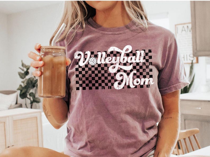 Puede incluir: Una camiseta malva con un gr&aacute;fico blanco que dice "Volleyball Mom" con un fondo a cuadros y un gr&aacute;fico de voleibol.