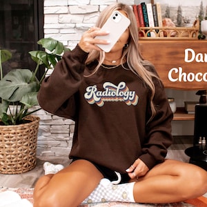 May include: Dark chocolate-colored sweatshirt with the word "Radiology" in a retro font. The text is in a rainbow color scheme. The sweatshirt is part of a clothing collection.