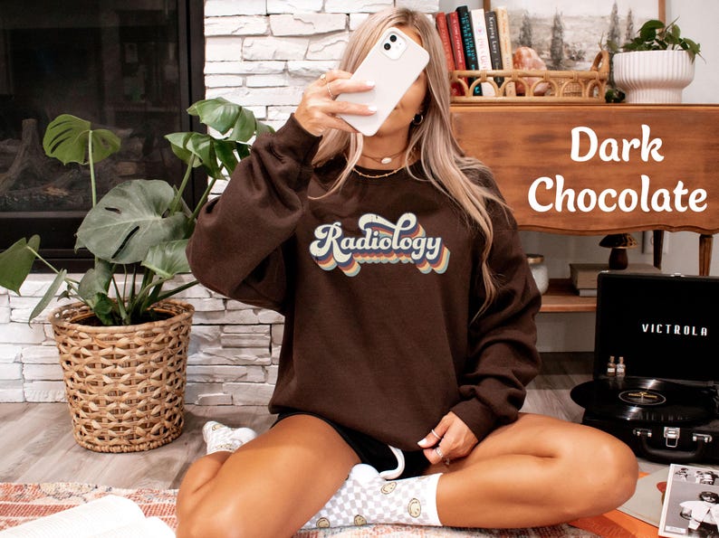 May include: Dark chocolate-colored sweatshirt with the word "Radiology" in a retro font. The text is in a rainbow color scheme. The sweatshirt is part of a clothing collection.