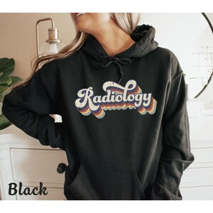 May include: Black hoodie with the word "Radiology" in a retro font, featuring a white outline and rainbow-colored accents. The hoodie has a drawstring hood and a front pocket. The word "Black" is printed at the bottom left.