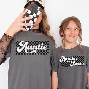 May include: Two gray t-shirts with the words "Auntie" and "Auntie's Bestie" in white script over a black and white checkered background. The shirts are a matching set, perfect for an aunt and niece or friend.
