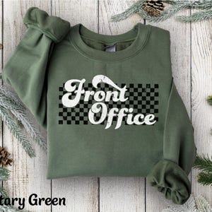 May include: Military green sweatshirt with the words "Front Office" in a white, retro font over a black and white checkered design. The sweatshirt has long sleeves and a crew neck.