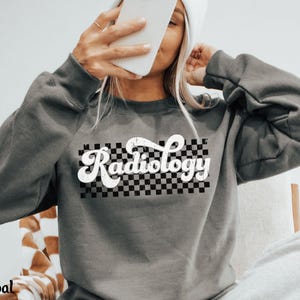 May include: Charcoal gray sweatshirt with the word "Radiology" in a white, retro font over a black and white checkered pattern. The sweatshirt has long sleeves and a crew neck.