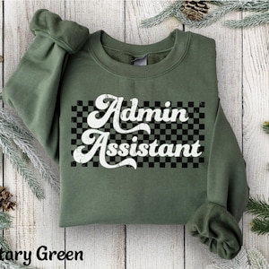 May include: Military green crewneck sweatshirt with the words "Admin Assistant" in a retro font over a black and white checkered pattern. The sweatshirt has long sleeves and a relaxed fit.