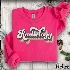 May include: A bright pink sweatshirt with the word "Radiology" in a retro font. The text is layered with a rainbow of colors, including blue, orange, and brown. The sweatshirt is a crew neck style.