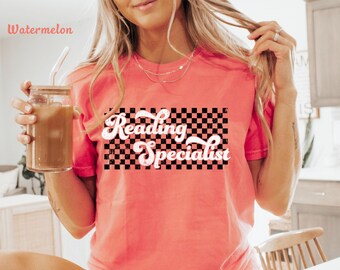 Op de afbeelding: Koraalrood T-shirt met de tekst "Reading Specialist" in een retro lettertype over een zwart-wit geblokt ontwerp. Het shirt is gemaakt van een zacht materiaal, perfect voor leraren.