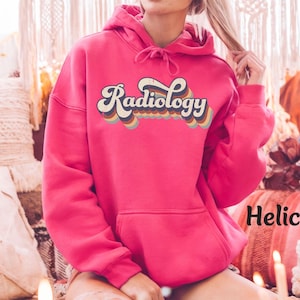 May include: A vibrant pink hoodie with the word "Radiology" in a retro font across the chest. The text is outlined in cream and features a rainbow color scheme. The hoodie has a drawstring hood and a front pocket.