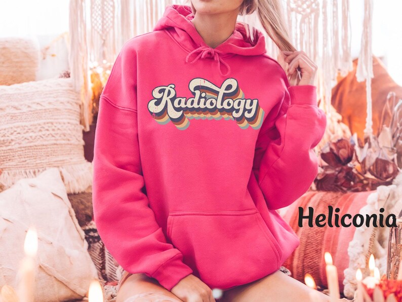May include: A vibrant pink hoodie with the word "Radiology" in a retro font across the chest. The text is outlined in cream and features a rainbow color scheme. The hoodie has a drawstring hood and a front pocket.