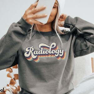 May include: Charcoal gray sweatshirt with the word "Radiology" in a retro font. The text is in white with a rainbow-colored outline. The sweatshirt has long sleeves and a crew neck. The word "Charcoal" is at the bottom left.