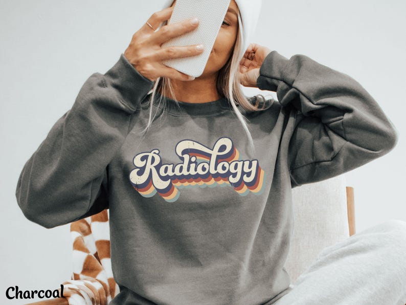 May include: Charcoal gray sweatshirt with the word "Radiology" in a retro font. The text is in white with a rainbow-colored outline. The sweatshirt has long sleeves and a crew neck. The word "Charcoal" is at the bottom left.