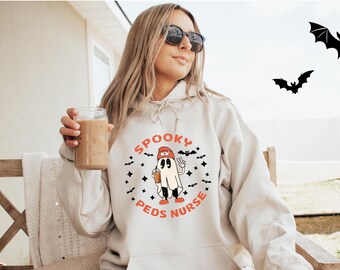 May include: A white hoodie with a graphic of a ghost wearing a red hat and holding a cup. The text "Spooky Peds Nurse" is printed in orange on the hoodie.