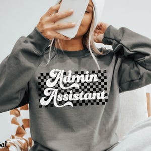 May include: Charcoal gray sweatshirt with the text "Admin Assistant" in a retro font over a black and white checkered pattern. The sweatshirt has long sleeves and a crew neck.