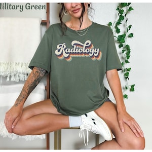 May include: Military green t-shirt with the word "Radiology" in a retro font. The text has a layered design with orange, yellow, and blue accents. The shirt is a short-sleeved, casual style.
