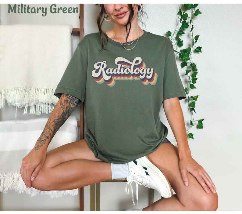 May include: Military green t-shirt with the word "Radiology" in a retro font. The text has a layered design with orange, yellow, and blue accents. The shirt is a short-sleeved, casual style.