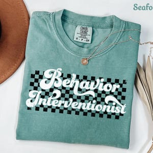 May include: Seafoam green t-shirt with a white checkered background and the text "Behavior Interventionist" in a retro font.