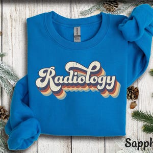 May include: Sapphire blue sweatshirt with the word "Radiology" in a retro font. The text has a layered design with orange, yellow, and blue stripes. The sweatshirt is displayed on a wooden surface with pine needles and pine cones.