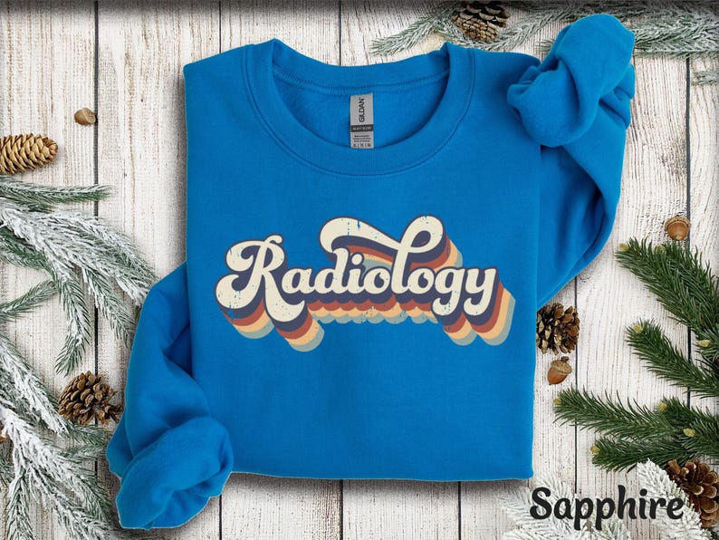 May include: Sapphire blue sweatshirt with the word "Radiology" in a retro font. The text has a layered design with orange, yellow, and blue stripes. The sweatshirt is displayed on a wooden surface with pine needles and pine cones.